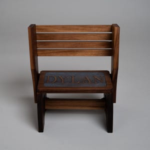 May include: A wooden chair with a slatted backrest and a rectangular seat featuring a dark gray inset panel. The name DYLAN is engraved in capital letters into the center of the seat panel. The chair is constructed from dark brown wood.