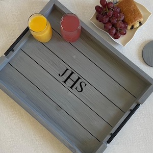 May include: A gray wooden serving tray with black handles and the initials "JHS" in black. Two glasses of juice, a plate of grapes and pastry, and a coaster are on the tray.