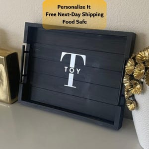 May include: A rectangular black wooden serving tray with side handles. The center of the tray features a large white letter T with the word TOY printed in smaller white letters underneath. The tray has a slatted base design.