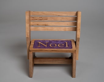 May include: A small wooden chair with a slatted backrest and a seat featuring a purple resin inlay. The word Noel is carved into the center of the purple seat in light-colored wooden lettering. The chair is constructed from light brown wood.