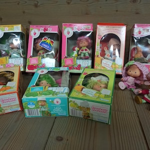 Może przedstawiać: Kolekcja lalek Strawberry Shortcake w stylu vintage w oryginalnych pudełkach. Pudełka są w jasnych kolorach z ilustracjami postaci i przezroczystymi plastikowymi okienkami. Jedna lalka jest poza pudełkiem.