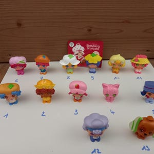 Peut inclure: Une collection de treize petites figurines en plastique Strawberry Shortcake, chacune avec des chapeaux et des tenues colorés uniques, disposées sur une surface blanche avec des chiffres bleus écrits à la main de un à treize et une carte logo rouge.