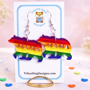 Puede incluir: Pendientes de oso de arcoíris con la palabra "MAMA" recortada. Los pendientes están en una tarjeta blanca con el texto "Tokyo Dog Designs" y un logotipo. Cuelgan de ganchos plateados.