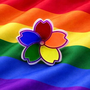 May include: A colorful flower-shaped pin with red, orange, yellow, green, and blue petals, outlined in white and purple. The pin rests on a rainbow flag, symbolizing LGBTQ+ pride and visibility.