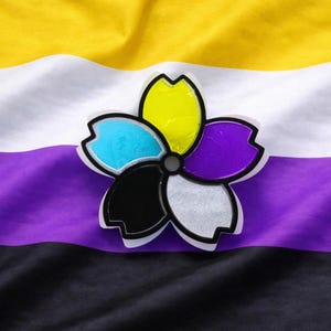 May include: A flower-shaped pin with petals in yellow, blue, black, silver, and purple, outlined in black. The pin is set against a non-binary pride flag background with yellow, white, purple, and black stripes. The pin is a symbol of identity and pride.