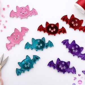 Puede incluir: Una colección de botones artesanales en forma de murciélago de colores rosa, rojo, turquesa y morado. Los botones tienen tres agujeros y están dispersos sobre una superficie blanca con cuentas rosas, lentejuelas y suministros de costura. También hay una lentejuela en forma de estrella.