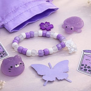 May include: A collection of purple accessories, including a beaded bracelet with white and purple beads, a drawstring bag, and stickers of a boba tea drink. Also included are a purple butterfly cutout, a flower, and a cat-shaped charm.