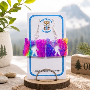 May include: A pair of dangle earrings shaped like the state of Oregon, featuring a vibrant, multi-coloured swirl pattern with glitter. The earrings are displayed on a white card with the text Tokyo Dog Designs and the website Tokyodogdesigns.com.