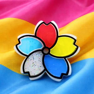 May include: A colorful flower-shaped pin with red, yellow, blue, and white petals, outlined in black. The pin is set against a background of pink, yellow, and blue fabric. The flower design is a brooch.