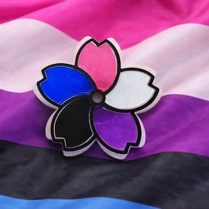 May include: A flower-shaped pin with petals in pink, blue, black, purple, and white, outlined in black. The pin is set against a draped background of pink, white, purple, black, and blue fabric, resembling a flag.