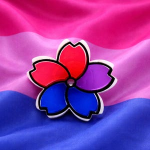 May include: A flower-shaped pin with red, purple, and blue petals, outlined in black and set against a background of pink, purple, and blue fabric. The pin has a white border and a small, clear gem in the center.
