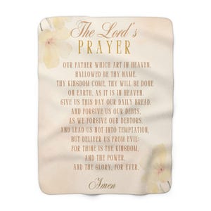 May include: A beige throw blanket with a watercolour floral design and the Lord's Prayer printed in brown text. The text reads: "The Lord's Prayer, Our Father which art in heaven, hallowed be thy name. Thy kingdom come, thy will be done on earth, as it is in heaven. Give us this day our daily bread. And forgive us our debts, as we forgive our debtors. And lead us not into temptation, but deliver us from evil: for thine is the kingdom, and the power, and the glory, for ever. Amen."