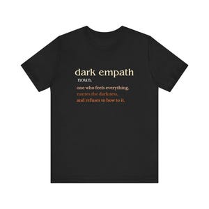 May include: Black t-shirt with the text "dark empath" in a serif font, followed by a definition. The definition reads: "one who feels everything, names the darkness, and refuses to bow to it."
