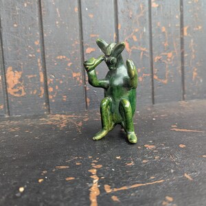 Op de afbeelding: Een kleine, groene keramische figuur van een zittend wezen met grote oren, die een klein kopje voor zijn gezicht houdt. Het beeldje heeft een glanzende afwerking en staat tegen een donkere, verweerde houten achtergrond.