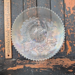 Puede incluir: Un plato de vidrio iridiscente de forma redonda con borde festoneado, que presenta un diseño en relieve de un pavo real y flores. Una regla de madera colocada verticalmente junto al plato indica un diámetro de unos 23 cm para el plato.