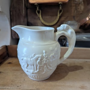 Może przedstawiać: Biały dzbanek ceramiczny z reliefową sceną polowania. Ucho ma kształt głowy psa.