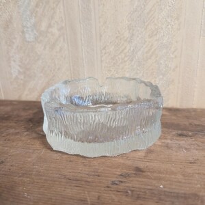 Puede incluir: Un cenicero de vidrio transparente y texturizado con un diseño circular. El cenicero tiene un exterior rugoso y desigual y un interior liso. El vidrio es grueso y parece vintage. El cenicero está sobre una superficie de madera.