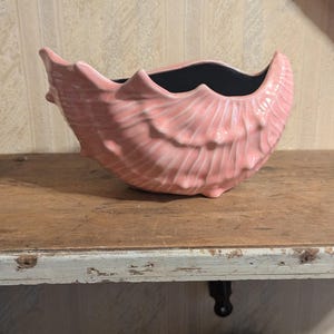 Może przedstawiać: Różowa ceramiczna miska w kształcie muszli z czarnym wnętrzem. Miska ma teksturowaną powierzchnię z żebrami i stoi na drewnianej półce. Półka ma postarzany, zużyty wygląd.
