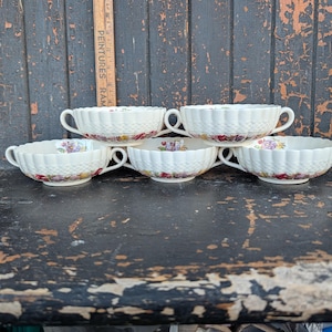 Peut inclure: Cinq bols à soupe en porcelaine blanche avec des anses. Chaque bol présente un motif côtelé et un motif floral sur le bord. Les bols sont disposés sur une surface sombre et patinée.