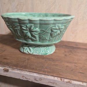 Puede incluir: Jardinera de cerámica verde menta con base de pedestal. La jardinera presenta un diseño detallado de hojas y vides alrededor del cuenco y la base. Colocada sobre una superficie de madera.