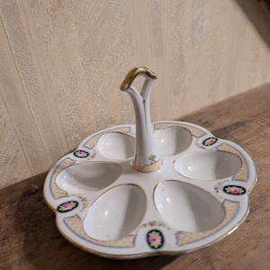 Puede incluir: Un servidor de huevos de porcelana blanca con seis hendiduras ovaladas y un asa central. El borde está decorado con un patrón de rejilla beige, detalles en azul claro y pequeños motivos florales en marcos ovalados negros con borde dorado.