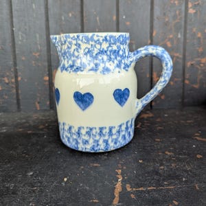 Puede incluir: Jarra de cerámica color crema con diseños de corazones y flores azules. La jarra tiene un asa y un pico. La parte superior e inferior de la jarra tienen un patrón floral azul. Tres corazones azules están centrados en la parte delantera.