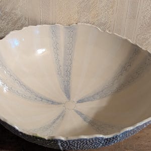 Pode incluir: Uma grande tigela de cerâmica feita à mão com uma borda exclusiva e ondulada. A tigela apresenta um interior creme com linhas azuis pontilhadas que irradiam do centro. A borda externa é azul texturizada, criando um contraste marcante.