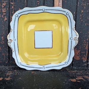 Może przedstawiać: Kwadratowy żółto-biały ceramiczny talerz do serwowania ze złotym obramowaniem. Talerz ma białą obwódkę z czarno-białym wzorem i złotymi akcentami. Środek talerza jest żółty z białym kwadratem pośrodku.