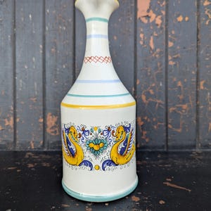 May include: A white ceramic vase with a flared top and a wide base. The vase features yellow, blue, and green painted details, including a decorative band with two yellow bird-like figures. The vase is approximately 25 cm tall.