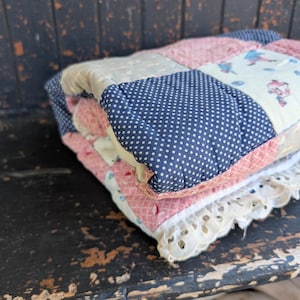 May include: A folded patchwork quilt with a navy blue polka dot pattern, pink and cream squares, and a white lace trim. The quilt is stacked on top of a white lace trimmed blanket.