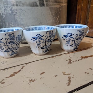 Puede incluir: Tres tazas de té blancas con diseños florales azules. Las tazas tienen una forma clásica con un borde acampanado y un asa. El patrón floral azul es intrincado y cubre el exterior de las tazas.