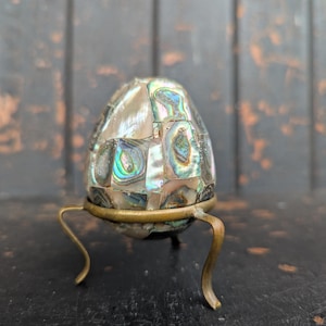 May include: An egg-shaped object made from abalone shell, displaying iridescent colours, mounted on a brass-coloured metal stand with three curved legs. The object is the focal point, showcasing a mosaic of shell pieces.