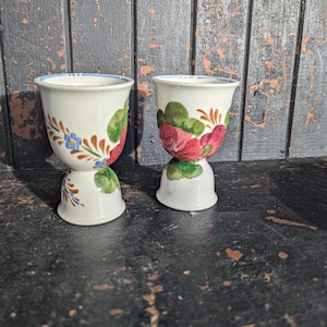 May include: Two white ceramic egg cups with floral designs. One cup features blue and orange flowers with green leaves, and the other has red roses and green leaves. Both have a blue rim and a pedestal base.