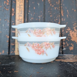 May include: Two stacked white milk glass bowls with side handles, featuring a horizontal grey band and a floral pattern of pink and peach blossoms with green leaves. A wooden ruler is visible behind the bowls, showing measurements in inches.