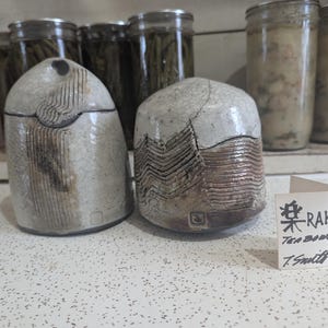 May include: Two ceramic tea bowls with a white glaze and brown and black markings. The bowls are sitting on a white countertop. A small white card with black writing reads "RAKU Tea Bowls T. Smith".