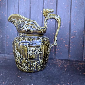 May include: An antique olive green ceramic jug with intricate relief detailing. The jug has a decorative handle with a horse's head, a spout with floral accents, and a detailed scene around the body. The jug is likely for decorative use.
