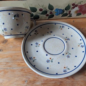 Pode incluir: Uma chávena de chá em cerâmica com pires a condizer, apresentando uma base de cor creme com um padrão pintado à mão de pequenas flores azuis e cor de laranja intercaladas com pontos azul-escuros. Ambas as peças têm um rebordo azul-escuro.