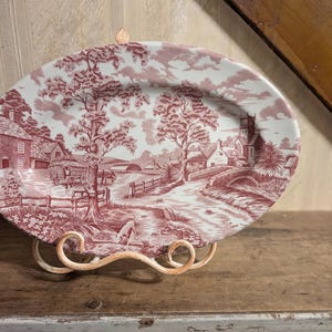 Może przedstawiać: Owalny półmisek ceramiczny z detalicznym, monochromatycznym nadrukiem w kolorze bordowym, przedstawiającym wiejski krajobraz. Scena ukazuje wioskę z domami, kamiennym mostem, drzewami oraz wozem konnym na ścieżce z małym psem.