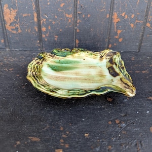 May include: A ceramic oyster-shaped dish with a textured, scalloped edge. The dish features a blend of green, tan, and brown glazes, creating a natural, earthy aesthetic. The dish is designed to resemble an oyster shell.