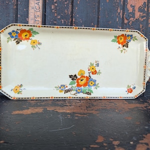 May include: A rectangular cream-coloured ceramic serving tray with scalloped handles and a floral pattern featuring orange, blue, and yellow flowers. The border has a black and orange geometric design. A wooden ruler shows a length of 30.5 cm.