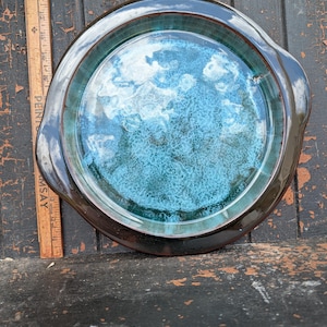 Può includere: Piatto da portata rotondo in ceramica con interno screziato blu e turchese e bordo marrone scuro. Il piatto ha due manici e una finitura lucida. Un righello di legno vintage è accanto al piatto.