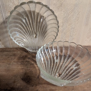 May include: Two clear glass seashell-shaped dishes. The dishes have a scalloped edge and a ribbed interior design. They are on a wooden surface. The dishes are likely for serving or display.