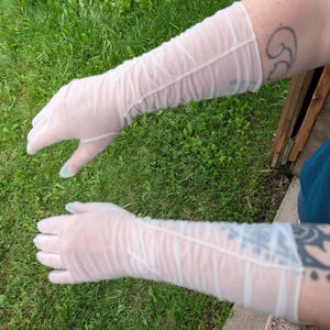 May include: A pair of sheer white opera-length gloves made of a lightweight, semi-transparent fabric. The gloves extend past the elbow and feature a gathered, ruched texture throughout the length of the sleeves, ending at the fingers.