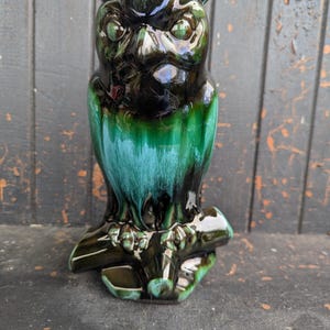 May include: A ceramic owl figurine with a glossy finish. The owl is predominantly green and black, with a teal-colored underbelly. It is perched on a branch, and the details of the owl's face and feathers are well-defined.