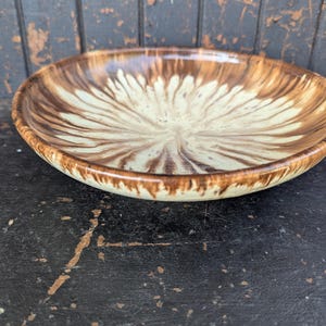 May include: A ceramic bowl with a brown and cream floral design. The bowl has a wide, shallow shape with a dark brown rim. The interior design features a cream centre with brown, petal-like strokes radiating outwards.