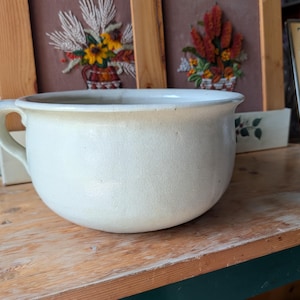 May include: A cream-colored ceramic chamber pot with a smooth, rounded body and a single side handle. The vessel features a wide, open top with a slightly flared rim and a solid, uniform off-white glaze finish across its entire exterior surface.
