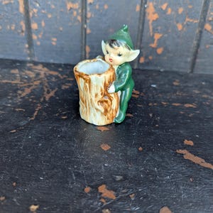Puede incluir: Pequeña figura de cerámica de un elfo con traje y sombrero verdes, sosteniendo un tocón de árbol en miniatura. El elfo tiene tez clara, cabello negro y está sonriendo. El tocón es marrón con un centro hueco.