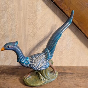 May include: A ceramic bird figurine featuring a long, sweeping tail and a slender neck. The bird is primarily finished in a glossy blue glaze with textured feather details, a bright orange beak, and a dark green base shaped like foliage.