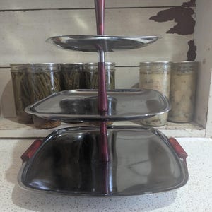 Può includere: Un vassoio da portata a tre livelli con manici in legno rosso e un montante centrale in legno rosso. Il vassoio è in metallo argentato e ha una finitura lucida.