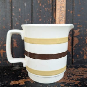 Peut inclure: Un pichet à crème en céramique blanche orné de rayures horizontales beige et marron foncé. Le pichet possède une anse carrée et un petit bec verseur. Une règle en bois est visible derrière, indiquant une hauteur d'environ 10 centimètres.